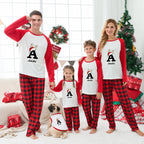 A family of four and a pug dog wearing matching red and black holiday pajamas with white sleeves, standing in a decorated living room with a Christmas tree, gifts, and a window behind them. Each person's pajama top has a large letter A with a reindeer graphic and the name Amelia printed below.
