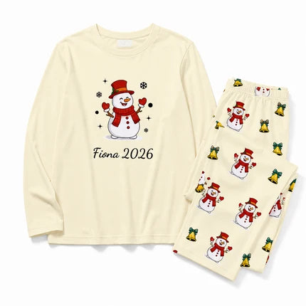 A two-piece cream-colored pajama set featuring a long-sleeve top with a cartoon snowman graphic and the text 'Fiona 2026', paired with matching pants printed with small snowmen and gold bells.
