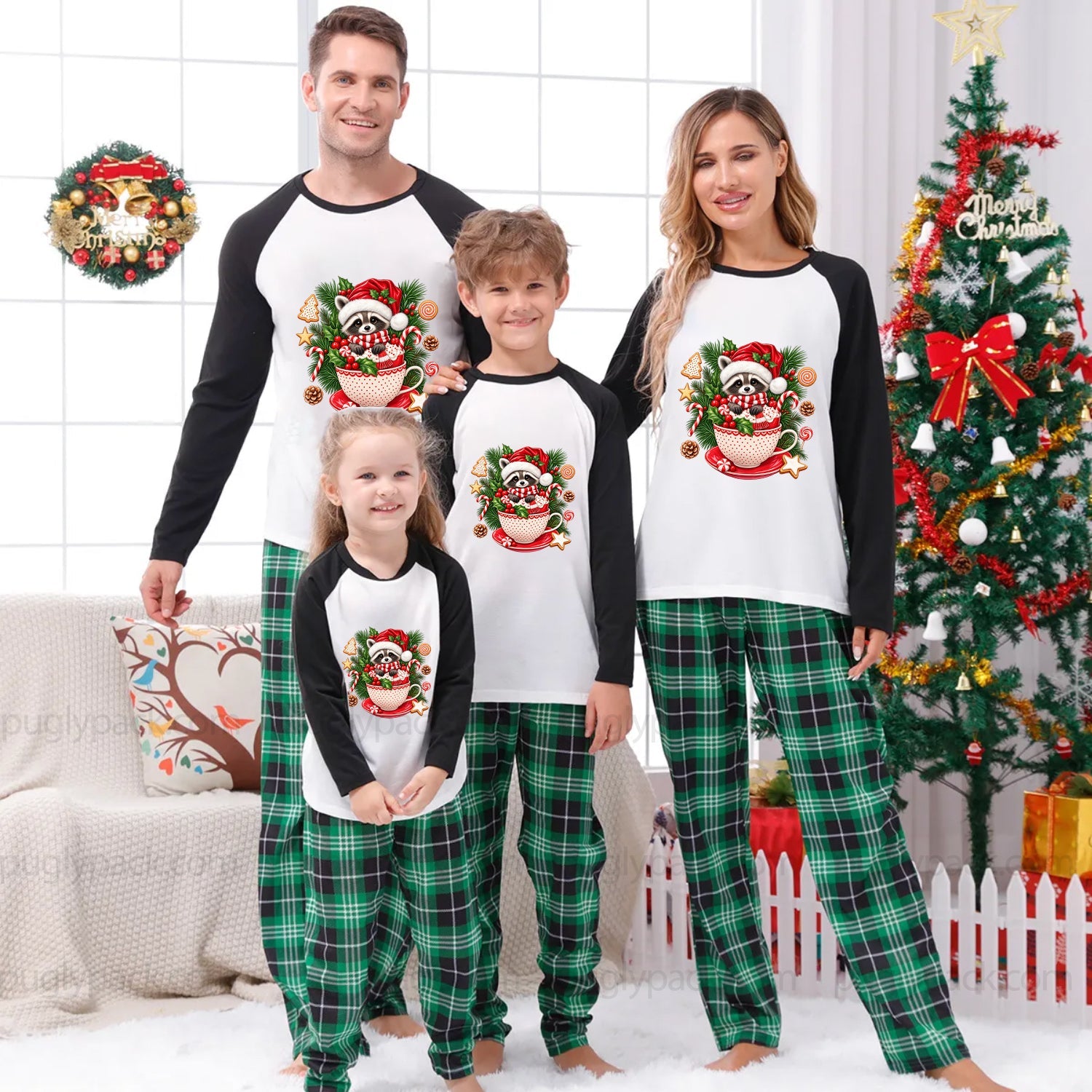 A family of four wearing matching Christmas raglan pajamas stands in a decorated living room with a Christmas tree, wreath, and holiday decor. The pajamas feature a white torso with black sleeves and a large Santa graphic, paired with green plaid pants.