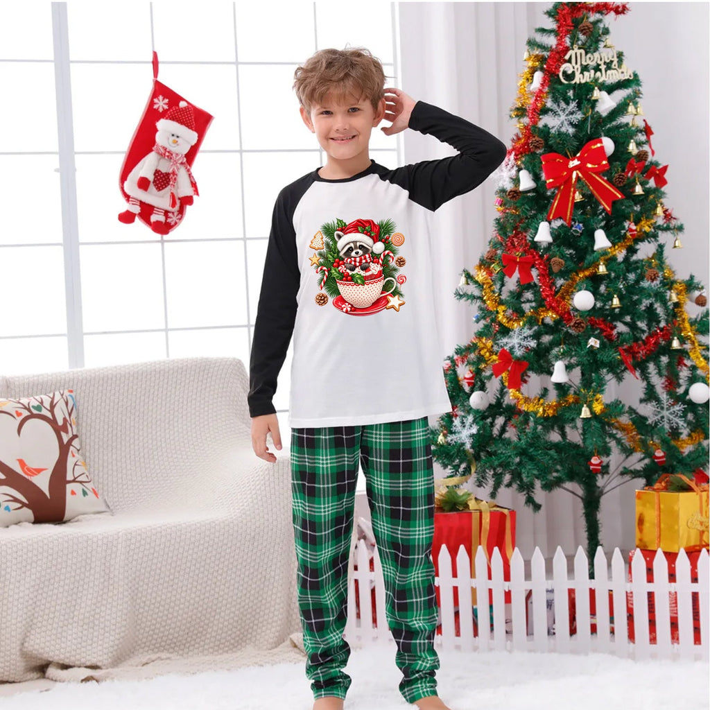 A young boy wearing a Christmas-themed raglan long-sleeve shirt and green plaid pajama pants stands in a decorated living room with a Christmas tree, stockings, and a couch.