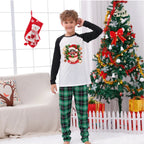 A young boy wearing a Christmas-themed raglan long-sleeve shirt and green plaid pajama pants stands in a decorated living room with a Christmas tree, stockings, and a couch.