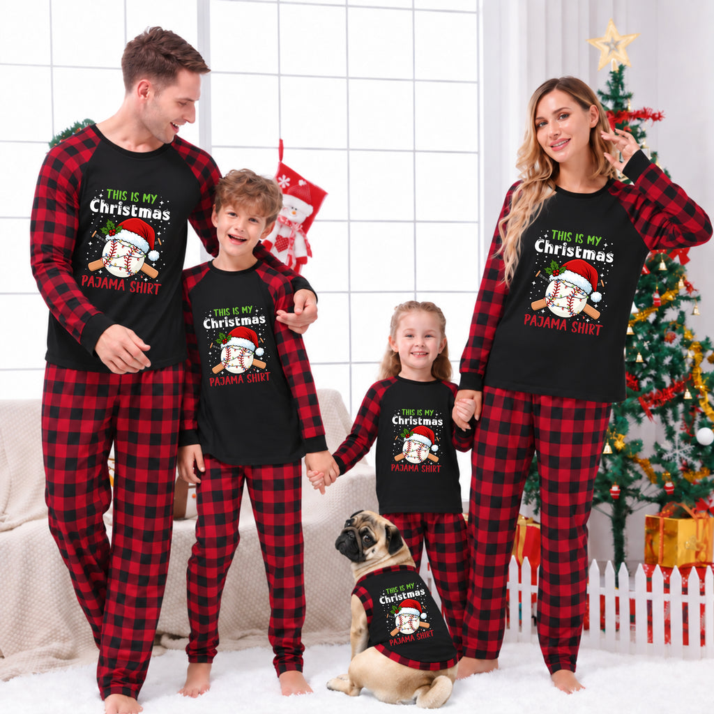 A family of four and a dog wearing matching red and black plaid Christmas pajamas with a black chest graphic that reads 'THIS IS MY Christmas PANDA SHIRT' in festive lettering. The background shows a decorated Christmas tree, stockings, and a cozy indoor holiday setting.