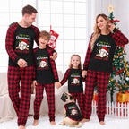 A family of four and a dog wearing matching red and black plaid Christmas pajamas with a black chest graphic that reads 'THIS IS MY Christmas PANDA SHIRT' in festive lettering. The background shows a decorated Christmas tree, stockings, and a cozy indoor holiday setting.