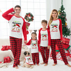 A family of four and a pug wearing matching Christmas pajama sets in a decorated living room. The pajamas feature a white and red raglan design with a cartoon penguin wearing a Santa hat and the text 'THIS IS MY Christmas PAMA SHIRT' above red and black buffalo plaid pants.