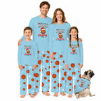 A family of four wearing matching light blue Christmas-themed pajamas with basketball and snowflake prints, standing together on a white background, with a small pug dog wearing a matching pajama set at the bottom right.